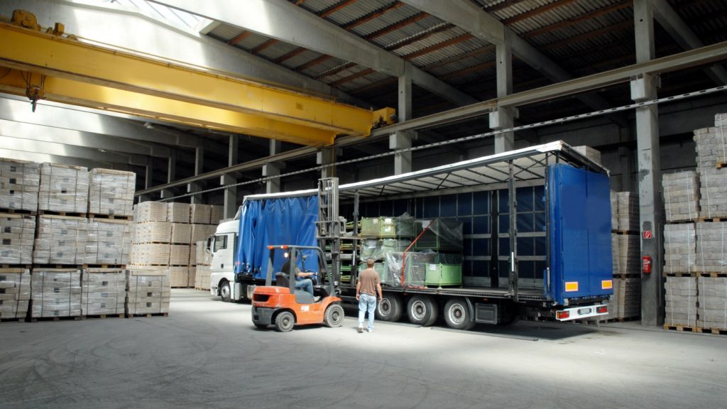 Ein LKW steht in einer Lagerhalle und wird von der Seite durch einen Gabelstabler und einer weiteren Person beladen